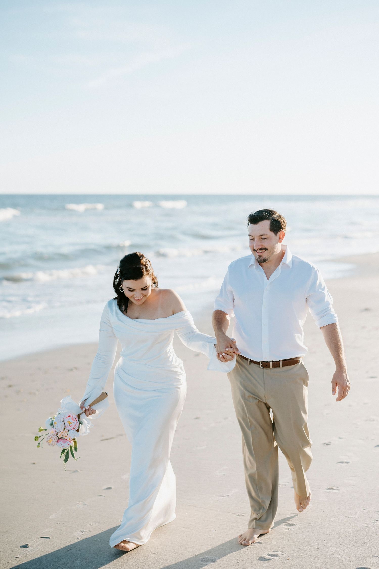 A couple walks hand in hand along a sunny beach shoreline wearing white and beige wedding attire.