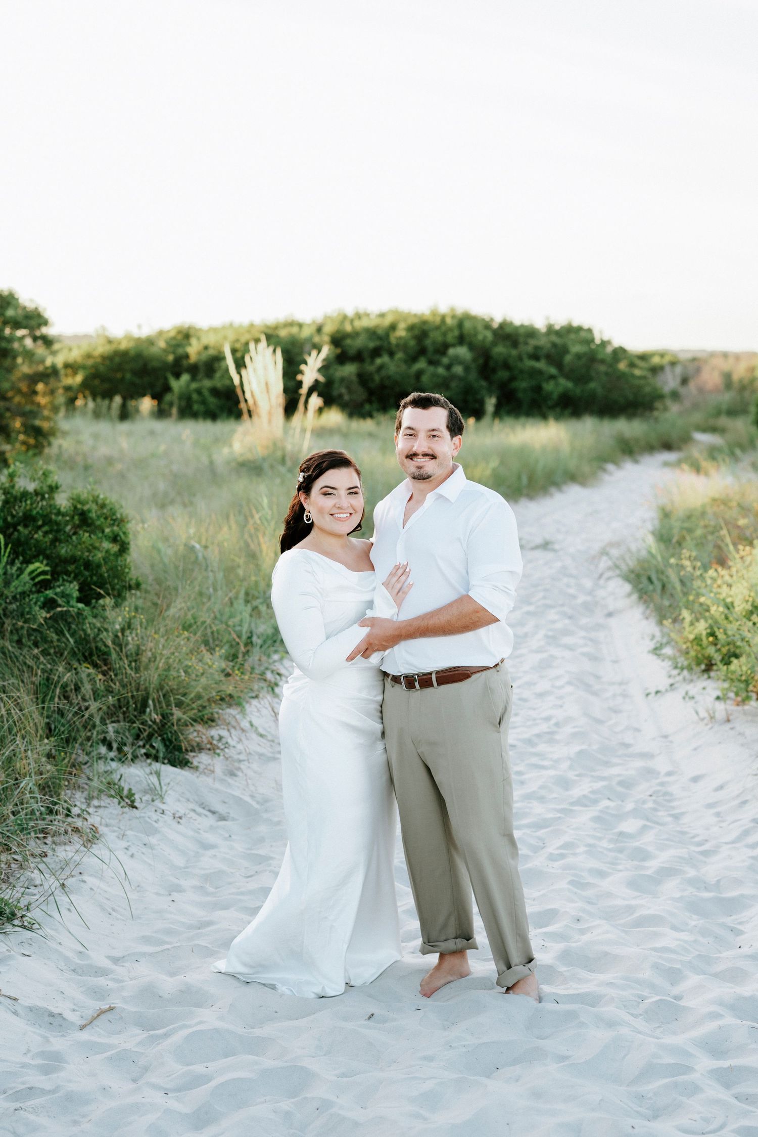 A couple shares a romantic moment on a sandy beach path surrounded by coastal vegetation at sunset.