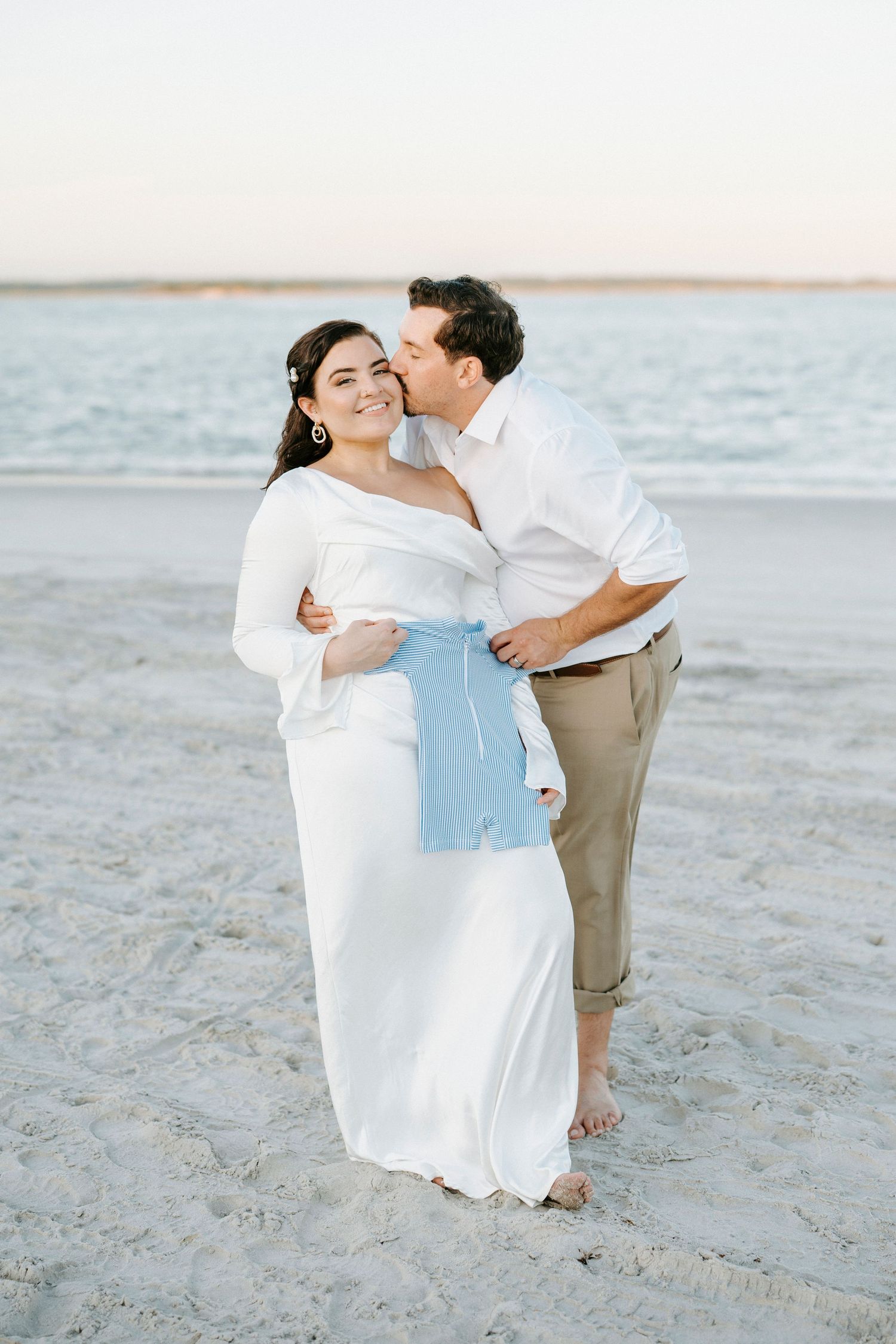 A couple shares romantic moments on a sandy beach at sunset wearing white and beige outfits.