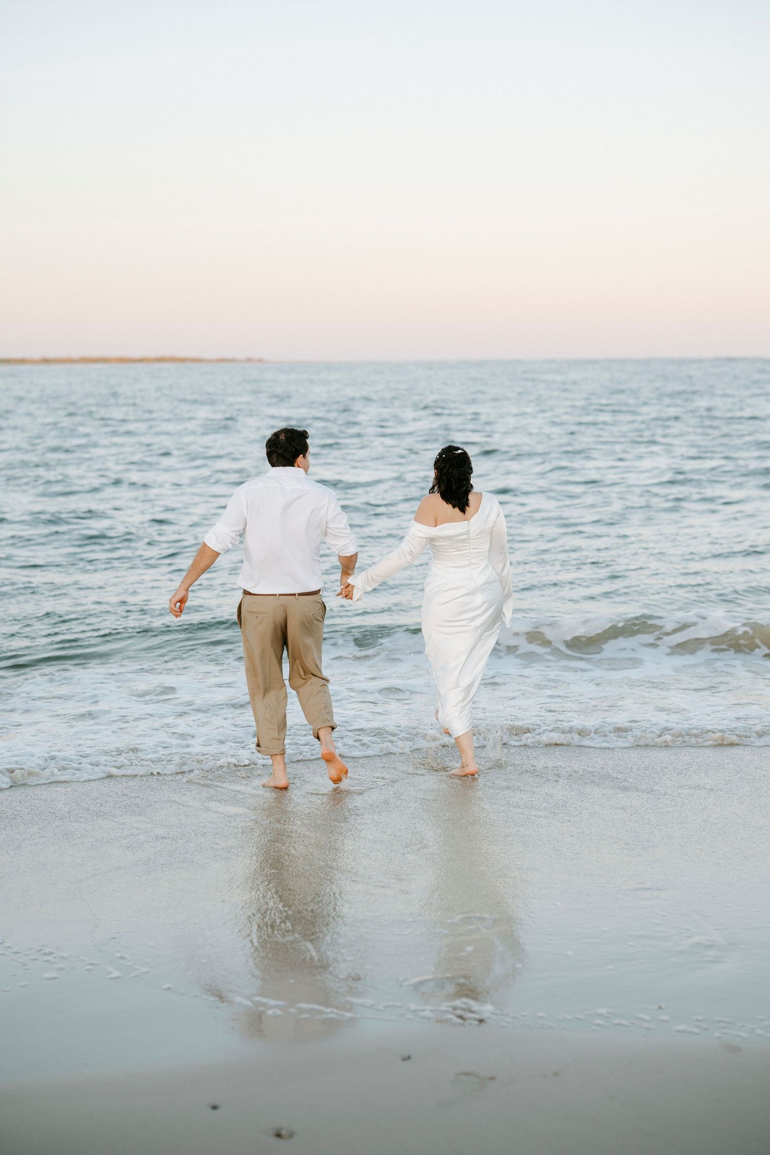 A couple playfully runs through ocean waves at sunset wearing white and beige casual outfits on the beach.
