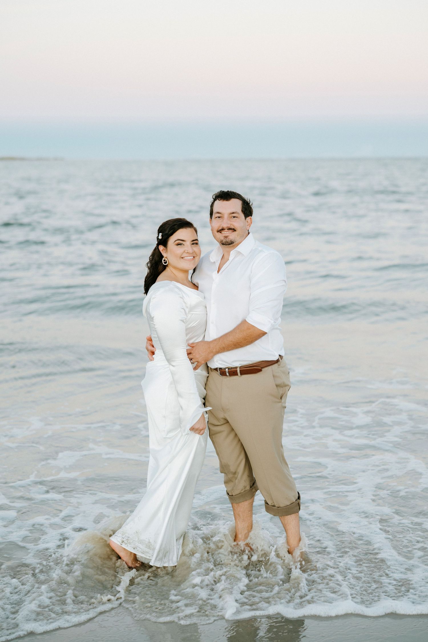 A romantic couple embraces while standing in shallow ocean water at the beach during sunset.