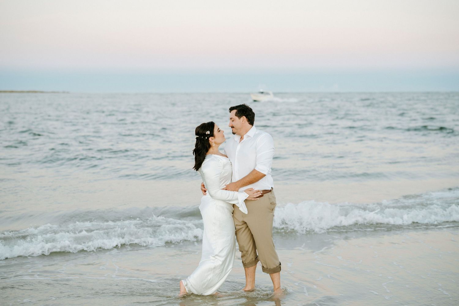 A couple in white and beige attire shares a romantic moment on a serene beach at sunset.