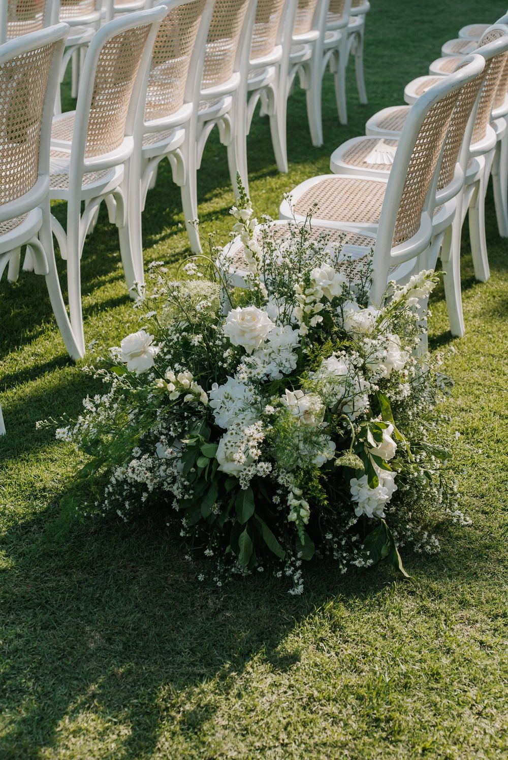 White floral arrangements line the grass aisle between rows of white chairs at an outdoor wedding ceremony.