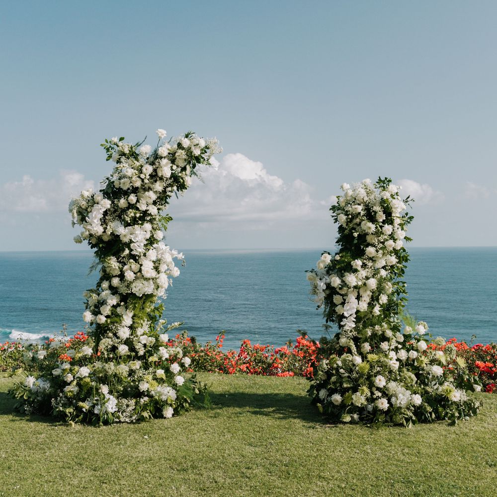 Two white floral wedding arches frame an ocean view on a grassy cliff overlooking blue waters and sky.