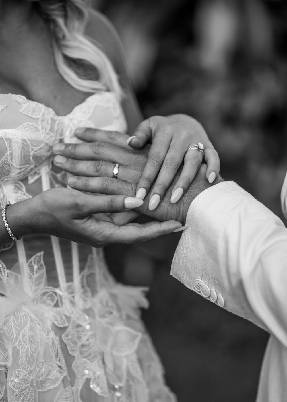 Black and white closeup of hands wearing wedding rings with lacy dress details visible in the background.