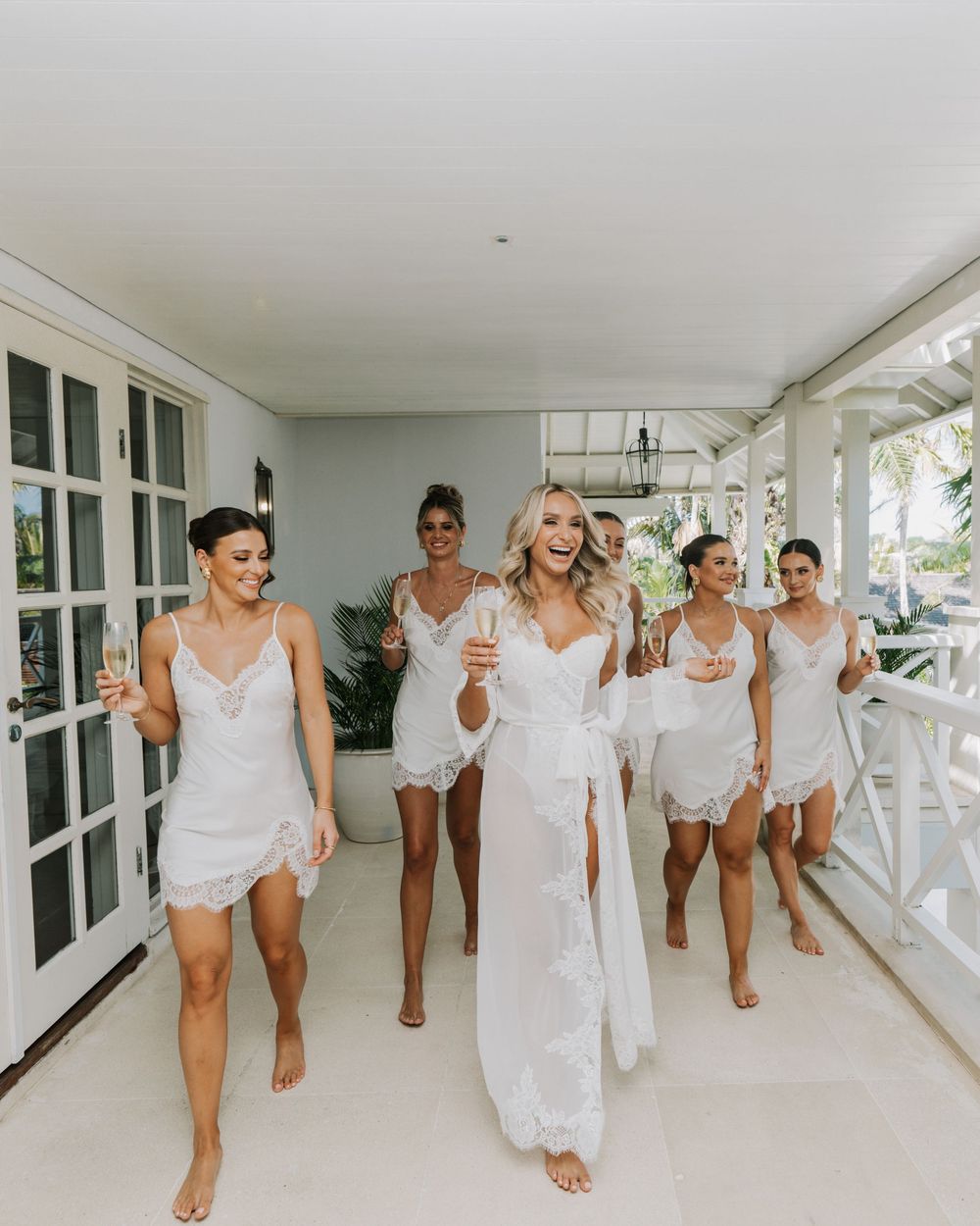 Bridal party in white outfits dancing and walking together through a bright covered porch walkway.