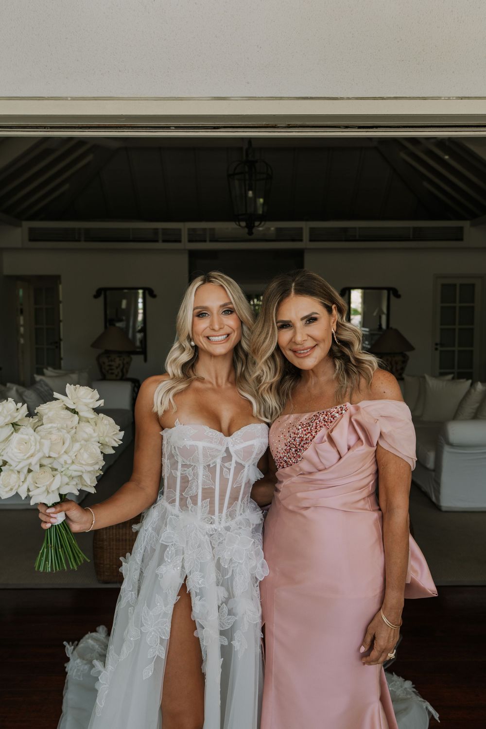 Two bridesmaids in elegant gowns, one in pink and one in white, smile while posing for wedding photos indoors.