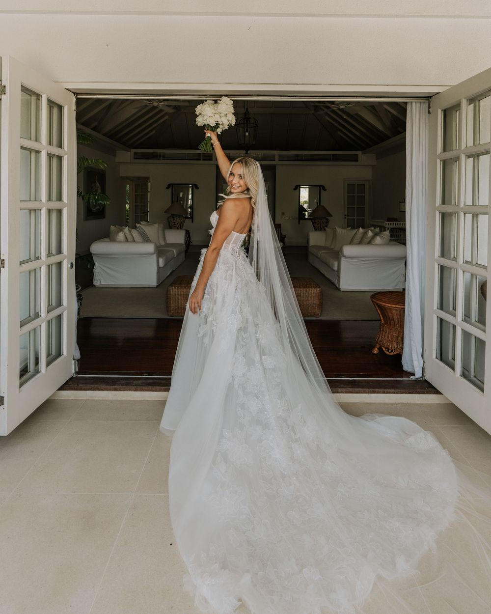 A bride in flowing white wedding gown with cathedral length veil glides across a bright sunlit room with French doors.