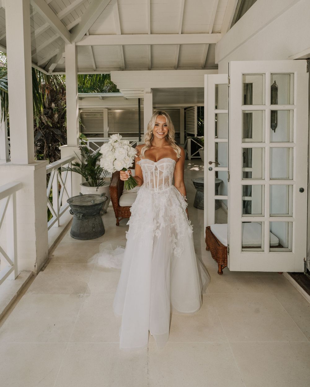 Elegant white wedding dress is displayed in a bright, modern room with French doors and indoor plants.