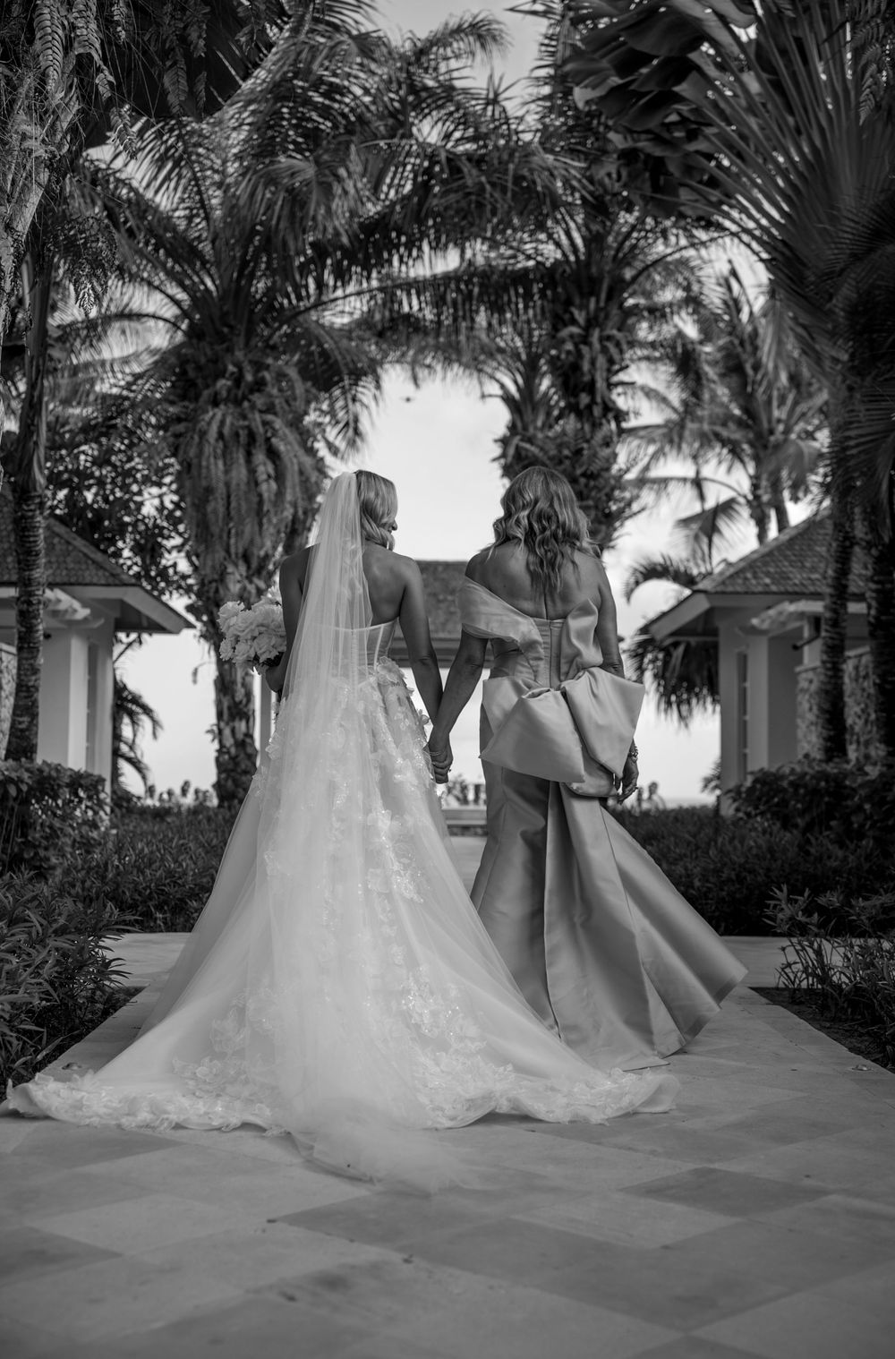 Two elegant wedding dresses photographed among tropical palm trees in a garden setting from different angles.
