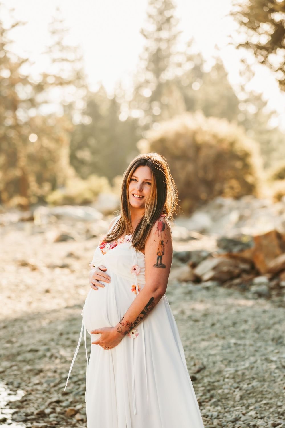 A pregnant woman in a flowing white dress stands in golden sunlight among rocks and trees at sunset.