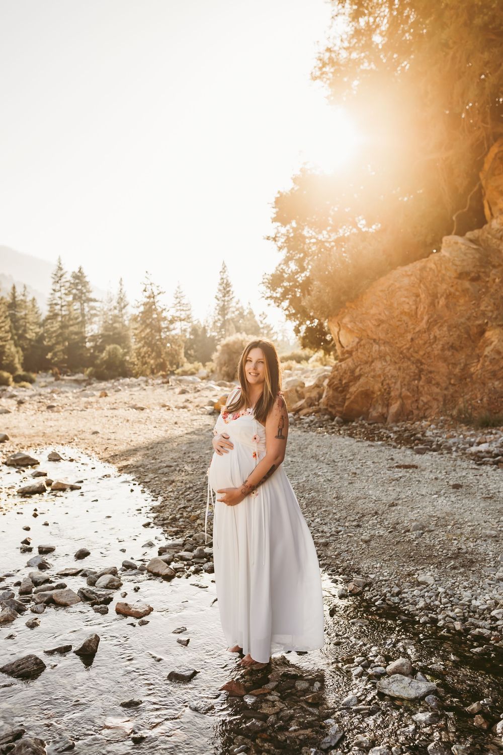 Maternity photo of woman in white dress standing at golden sunset on rocky river bank with tall pine trees in background.