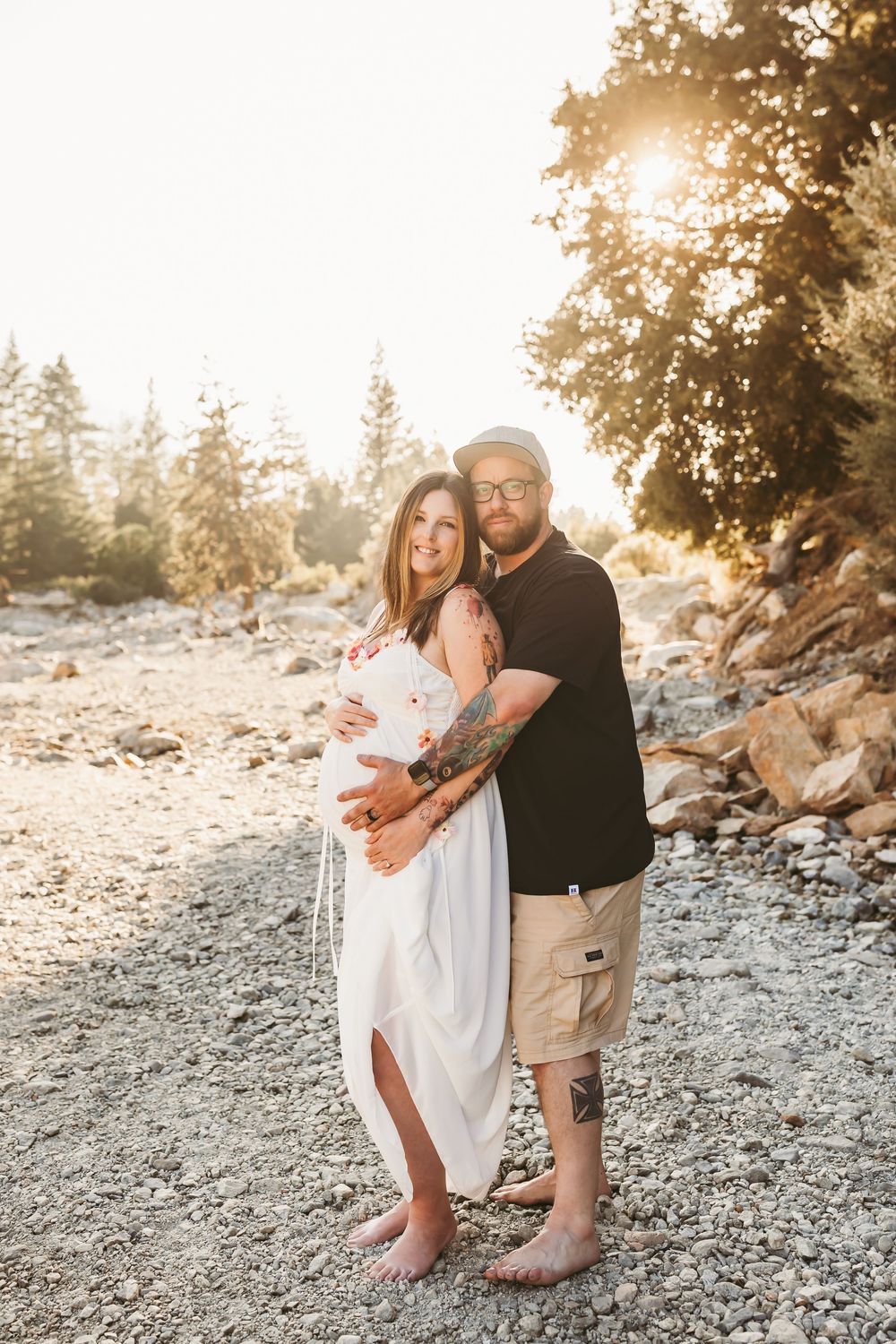 A couple embraces on a rocky beach during golden hour, creating a romantic silhouette against the sunlit trees.