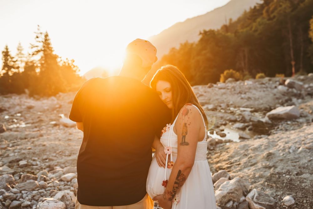 Two figures embrace during a golden sunset on a mountain beach with pine trees silhouetted in the background.