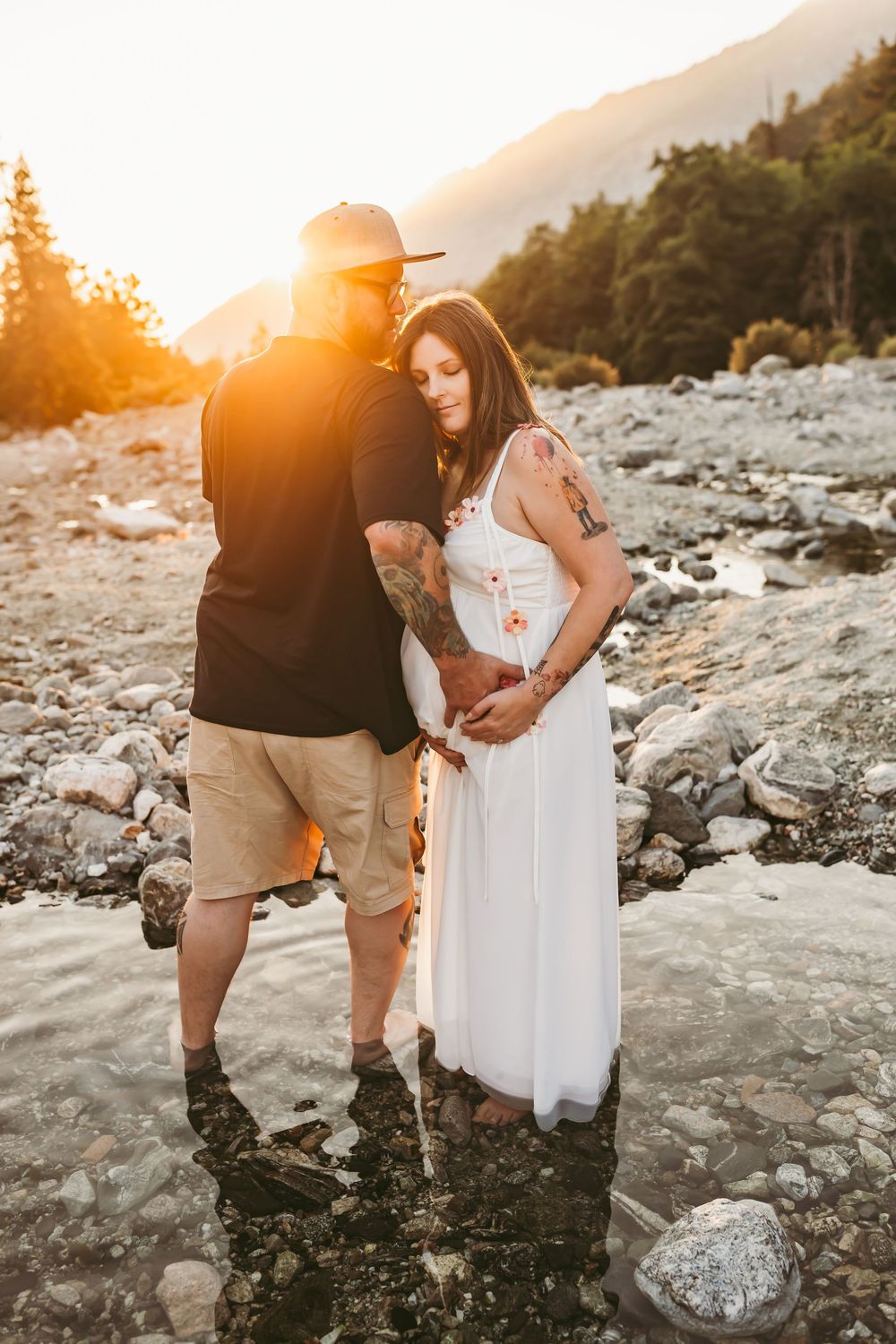 A couple shares an intimate moment on a rocky riverbank at sunset, with mountains and forest in the background.