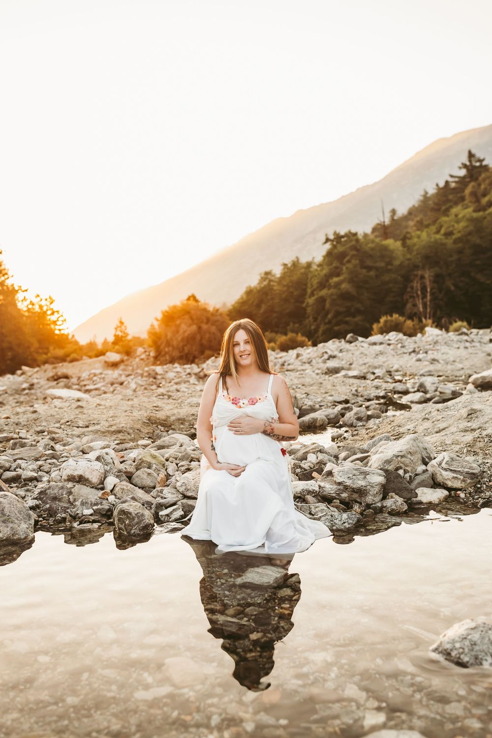 A woman in a flowing white dress sits gracefully on rocky coastal terrain at sunset, creating a dreamy beach portrait.