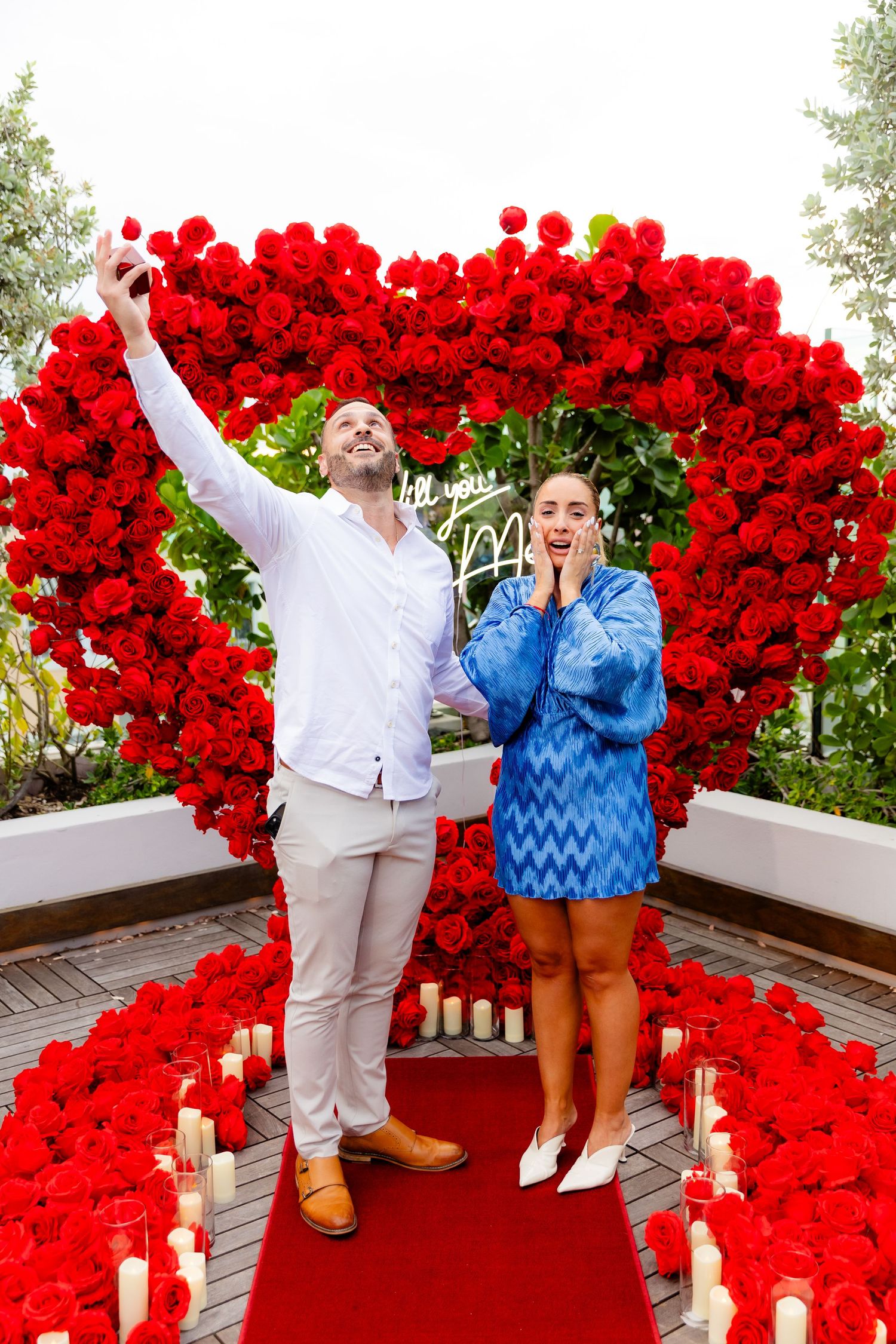 Vibrant red heart-shaped rose display with candles on wooden deck creates romantic outdoor atmosphere.