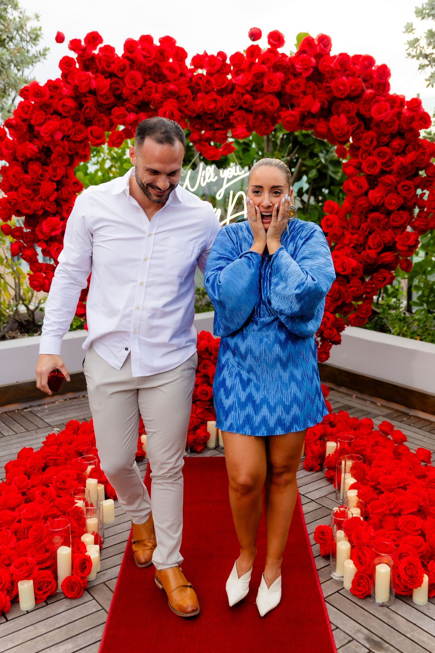 Two people celebrating in front of a romantic red rose heart-shaped arch on a wooden deck.