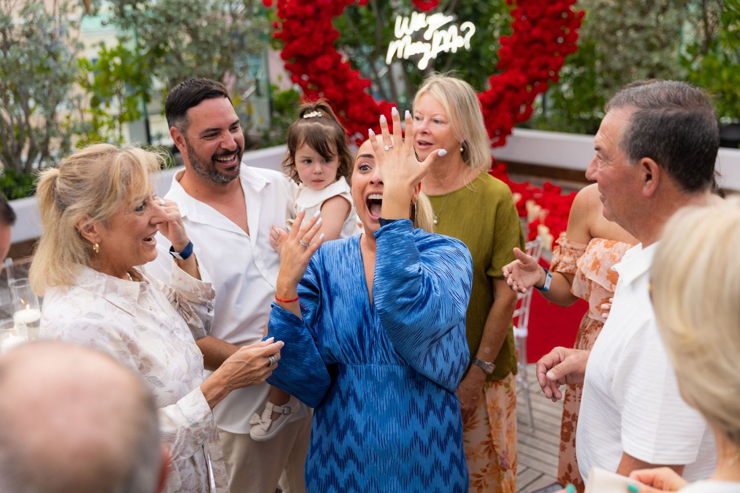 A surprise engagement moment captured under a stunning red rose heart arch with onlookers celebrating.