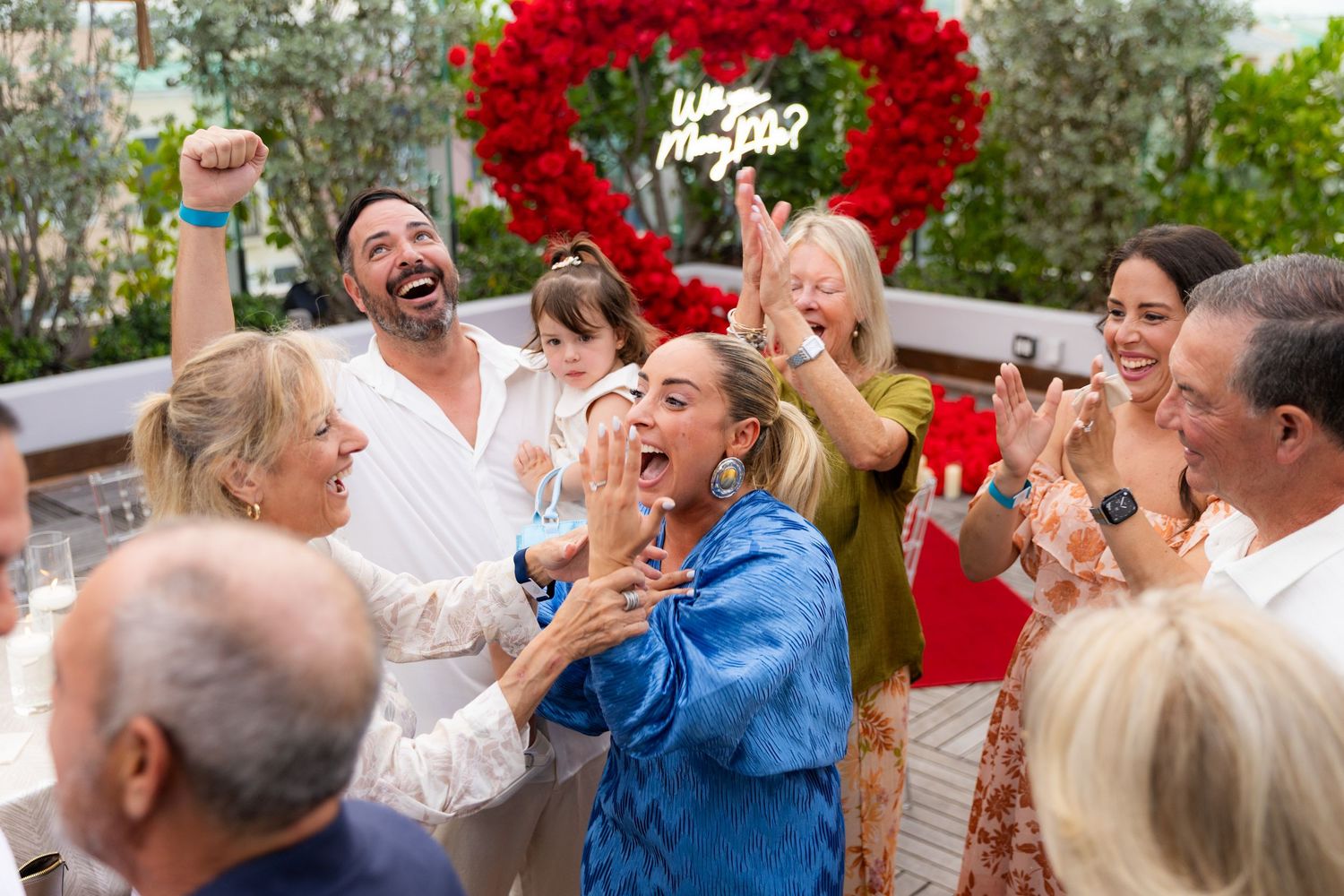 Friends and family react with joy during a marriage proposal under a red rose heart archway.