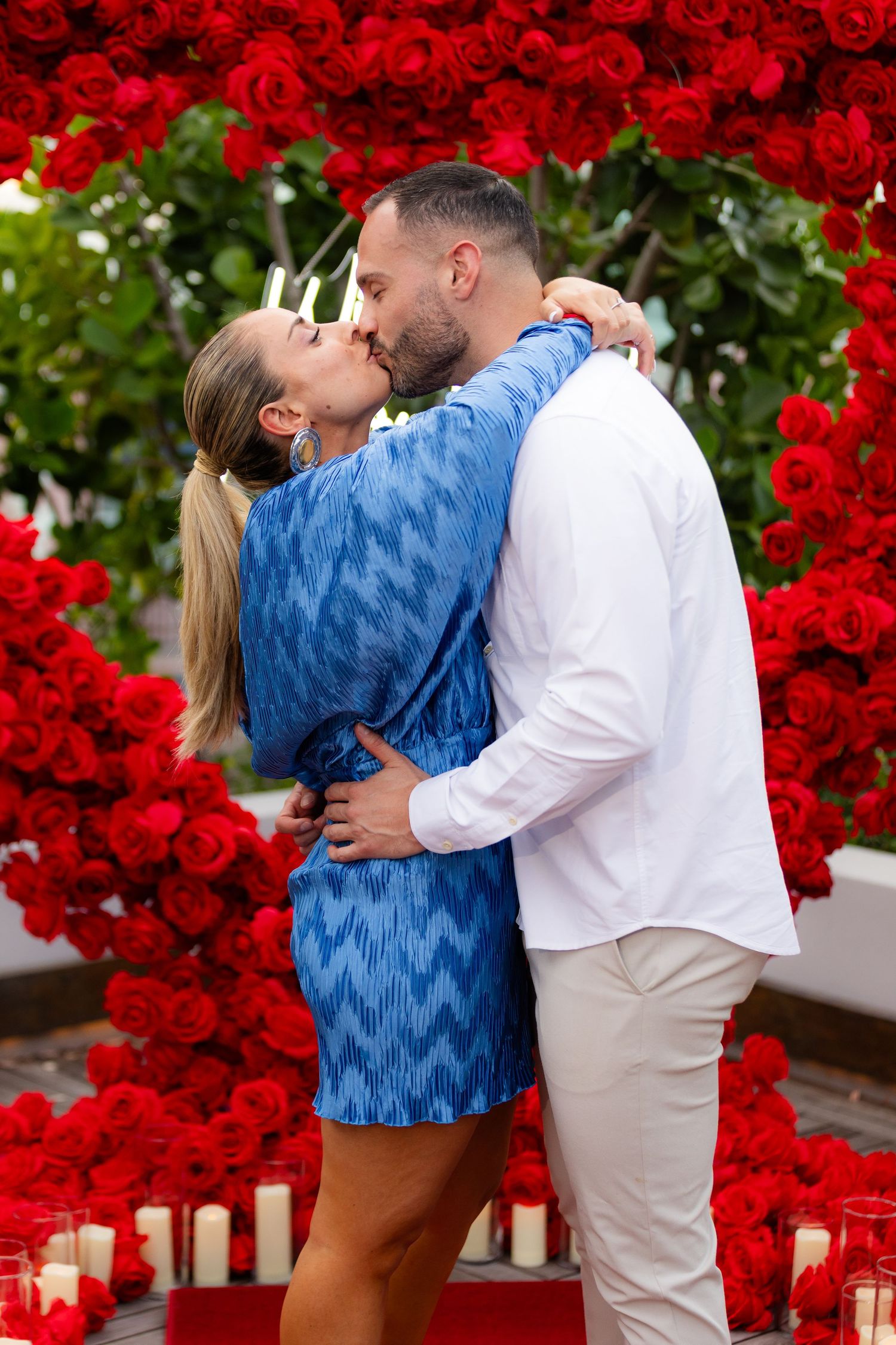 Couple embracing in front of a vibrant red rose wall with glowing candles below.