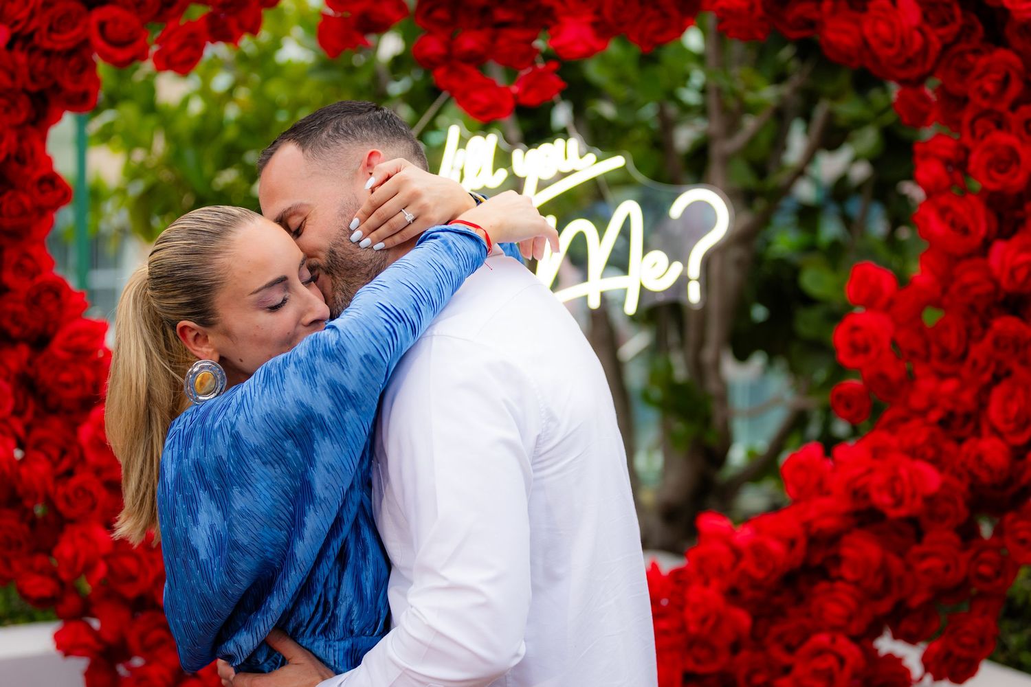 A couple embraces romantically beneath an archway of bright red roses during a special moment.