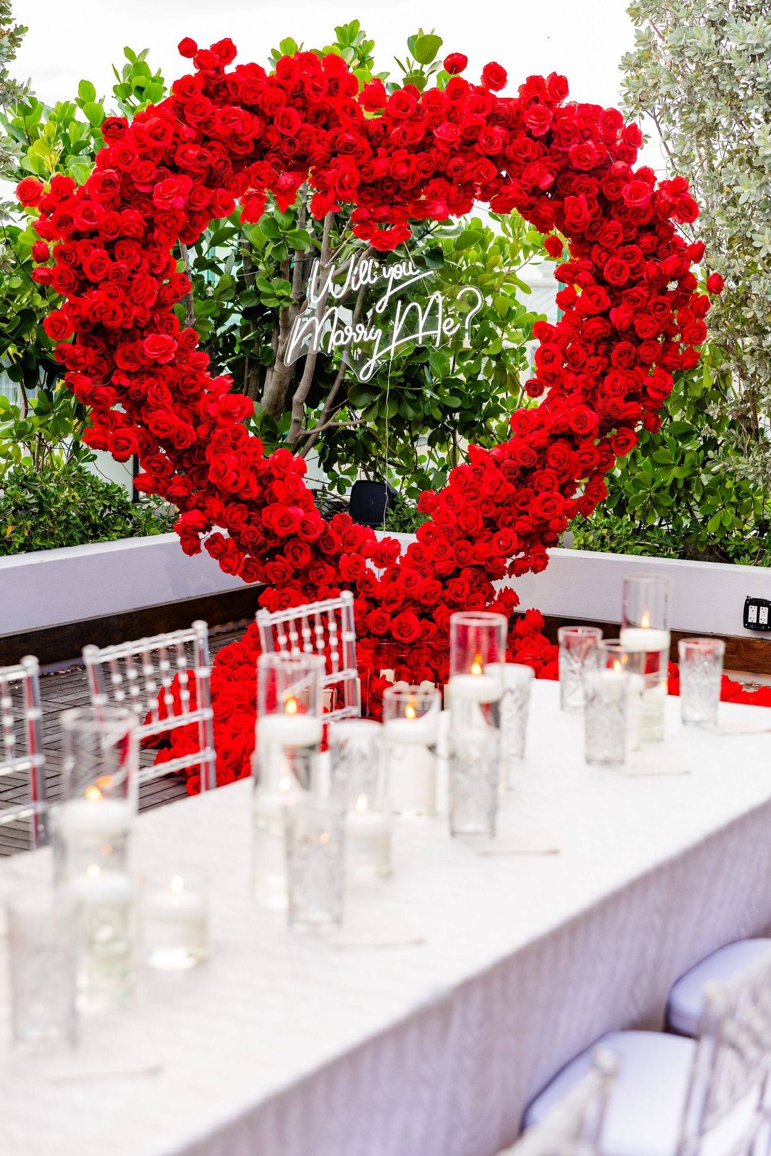 Romantic red rose arch with candles arranged on a white table for an outdoor event setting.
