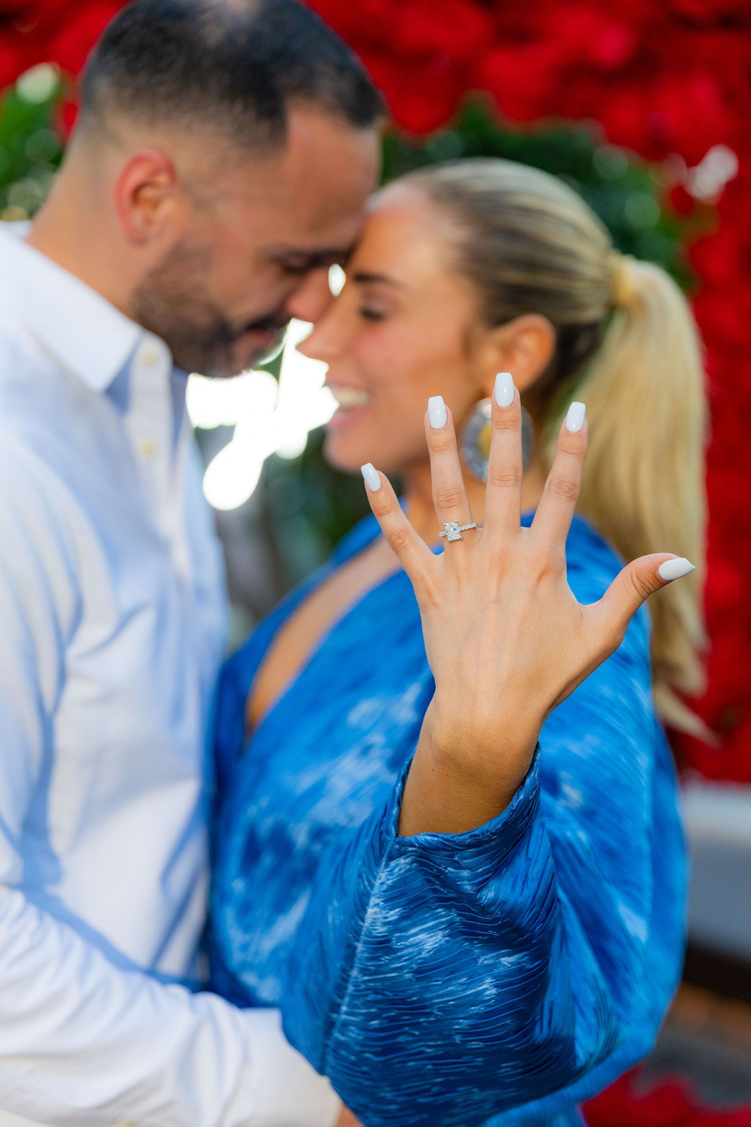 Couple embraces under arch of red roses while showing off engagement ring.