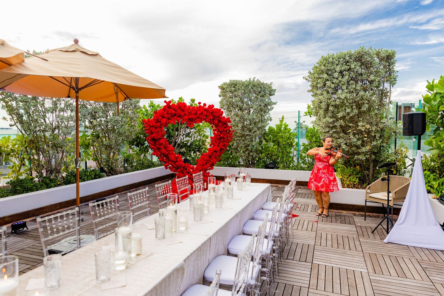Outdoor rooftop wedding venue setup with clear chairs and red heart-shaped floral arrangement under orange umbrella.