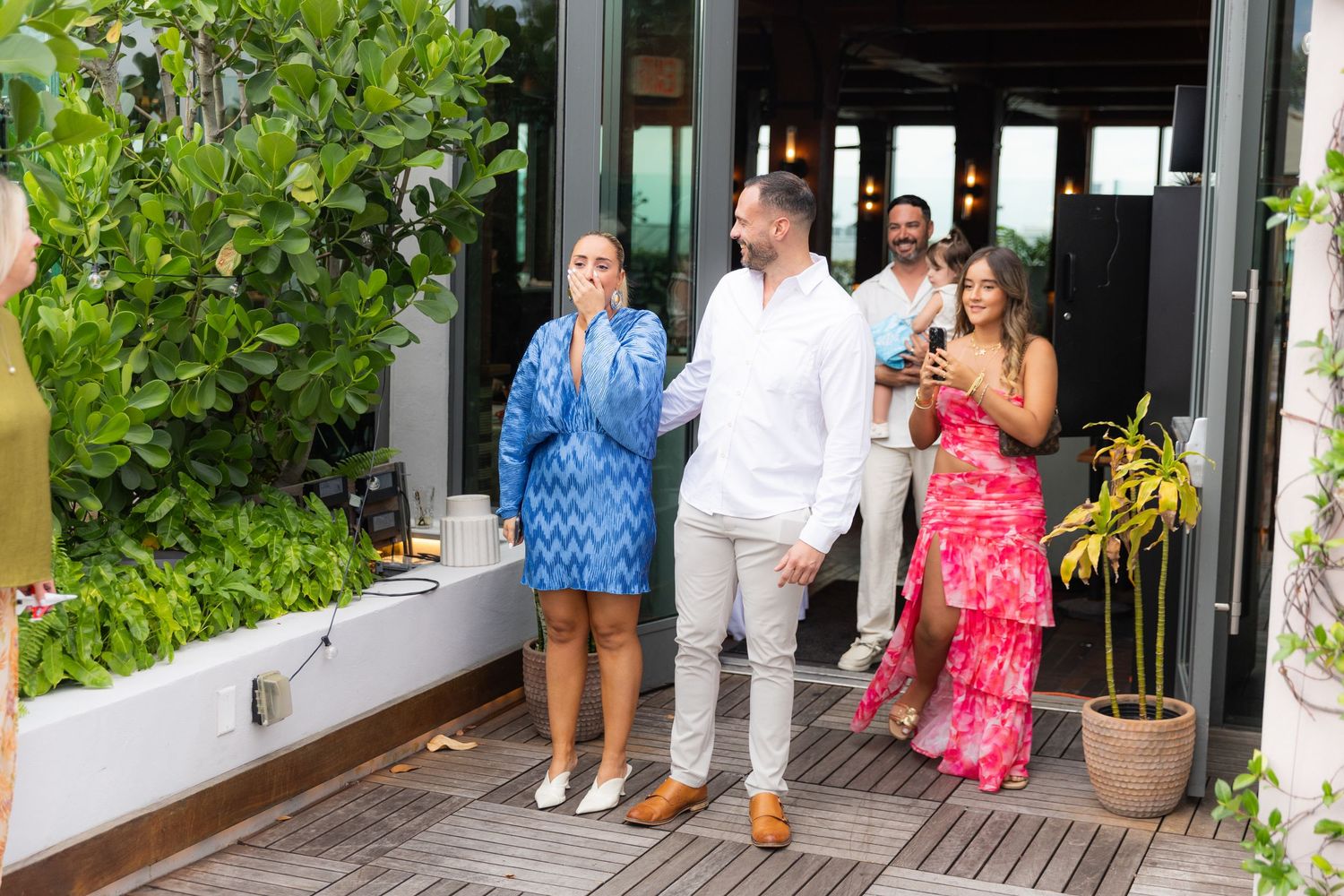 Event guests gather on a wooden deck outside a venue surrounded by lush green foliage.