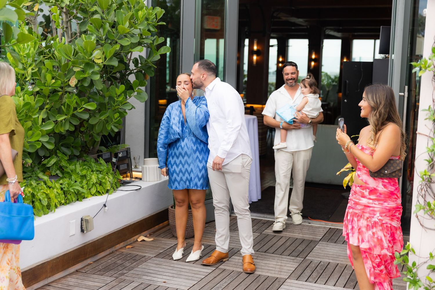 A wedding guest group gathers on a wooden deck patio near tropical greenery during a daytime celebration.