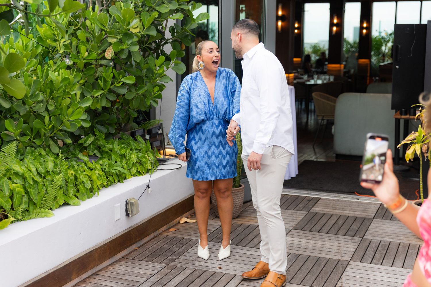 Two people stand on an outdoor wooden deck near green plants outside a restaurant.