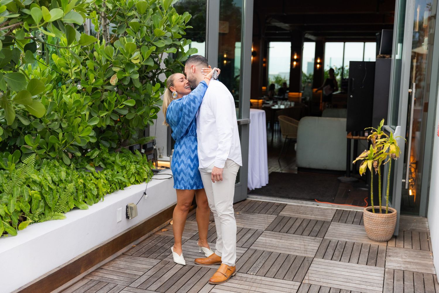 A couple shares a romantic moment on an outdoor deck with tropical plants and restaurant seating visible.