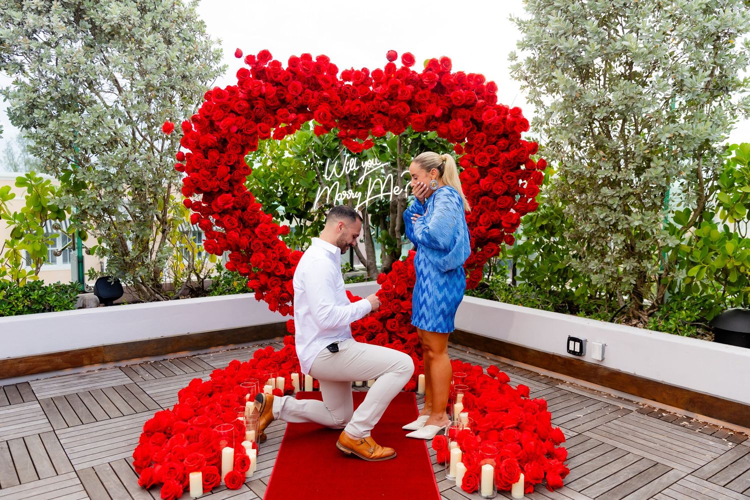 Intimate outdoor proposal setting with a dramatic red heart balloon installation against green foliage.