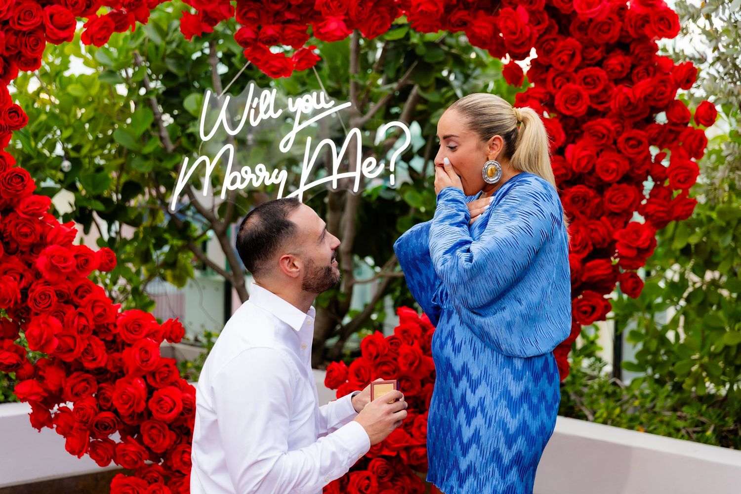 An elegant outdoor proposal scene with a giant red heart balloon arch and decorative text.
