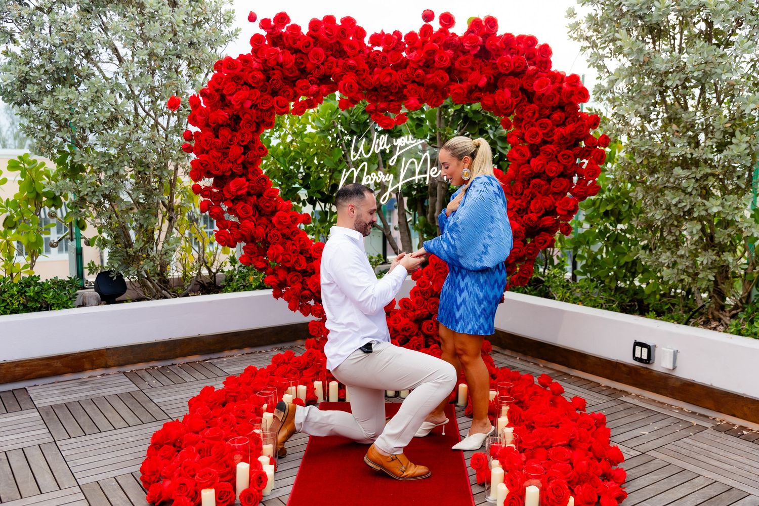 A large red balloon arch creates a romantic backdrop on an outdoor wooden deck surrounded by greenery.