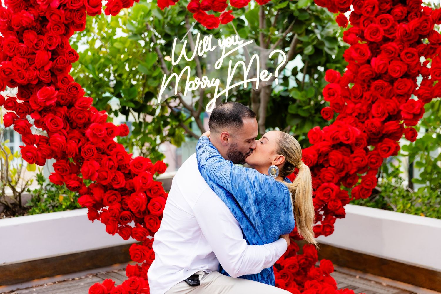 An elegant proposal scene with a giant red rose heart arch and candles on a wooden deck.