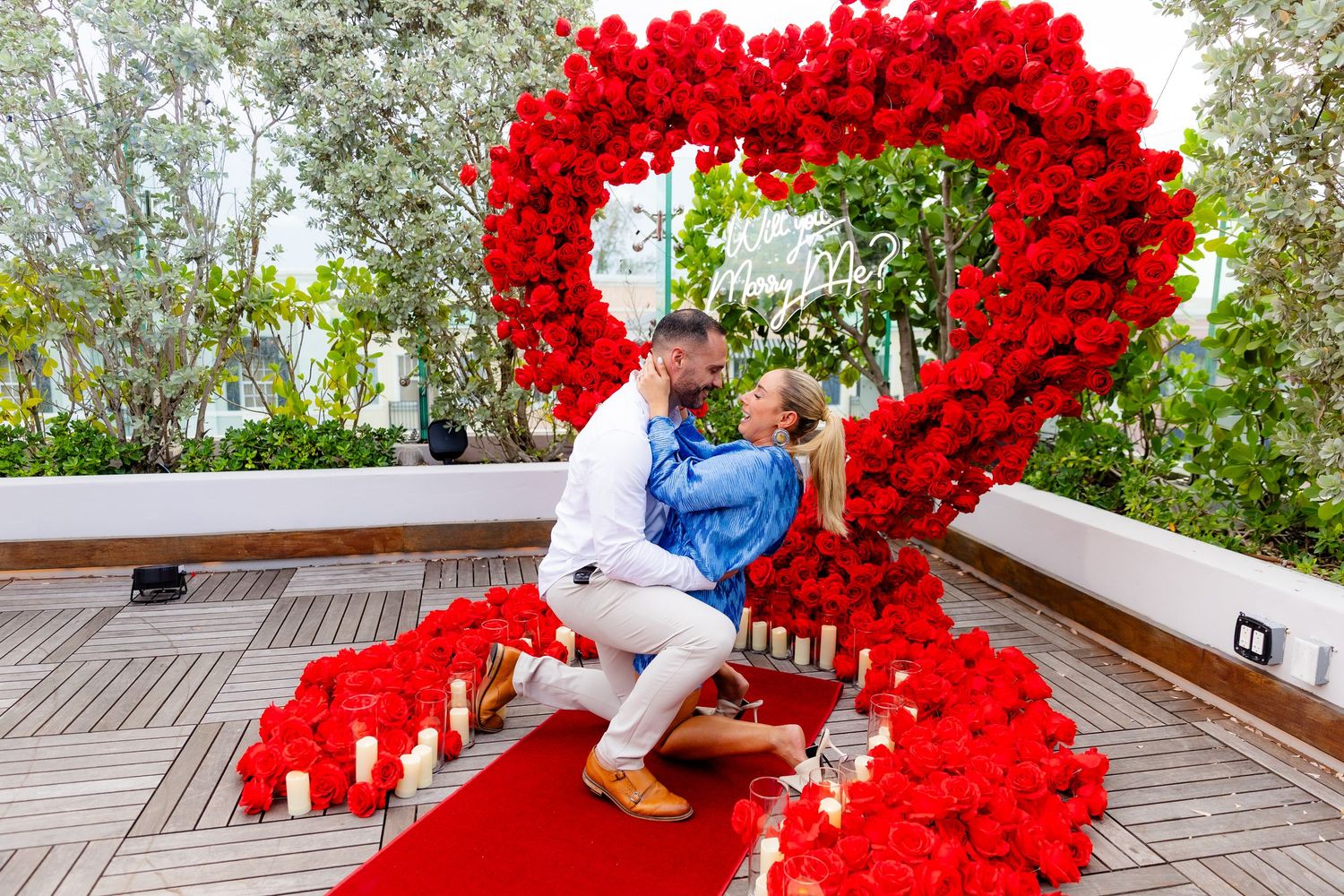Person sitting beside beautiful heart-shaped floral arrangement of red roses on outdoor wooden deck.