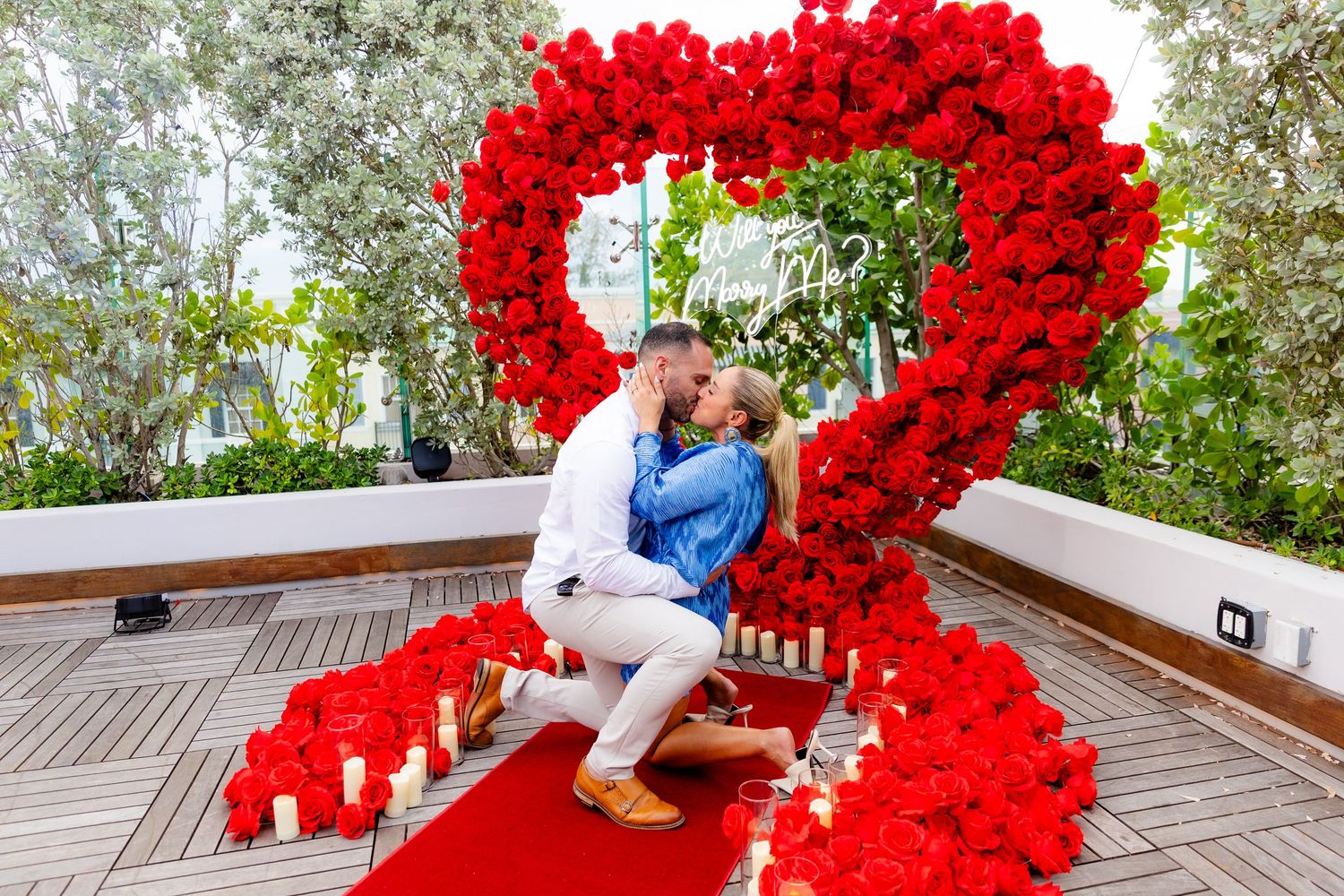 Large heart-shaped display made of bright red roses on wooden deck surrounded by lush greenery creates romantic scene.