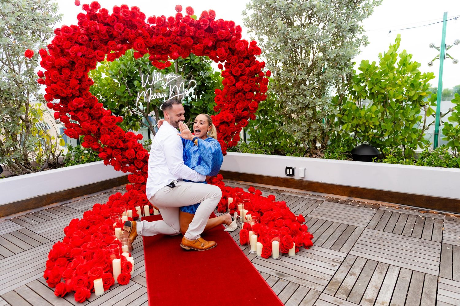Red heart-shaped balloon display decorates outdoor wooden deck with trees in background.