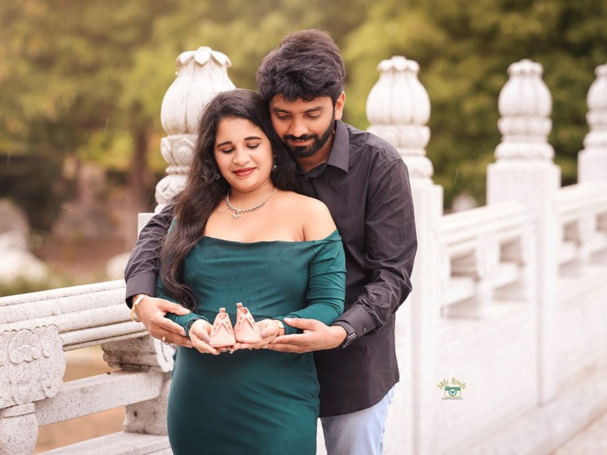 Couple poses in elegant maternity photo with navy blue gown against dark backdrop with cherry blossoms and water reflection.