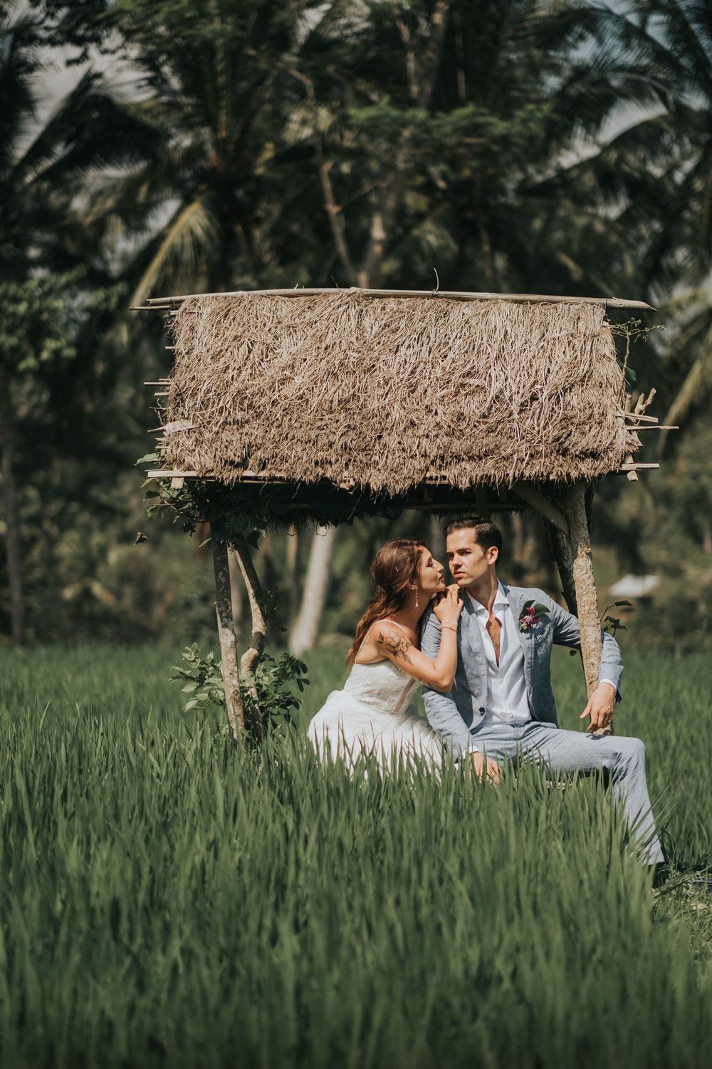 Couple sits under thatched roof shelter in lush green rice field.