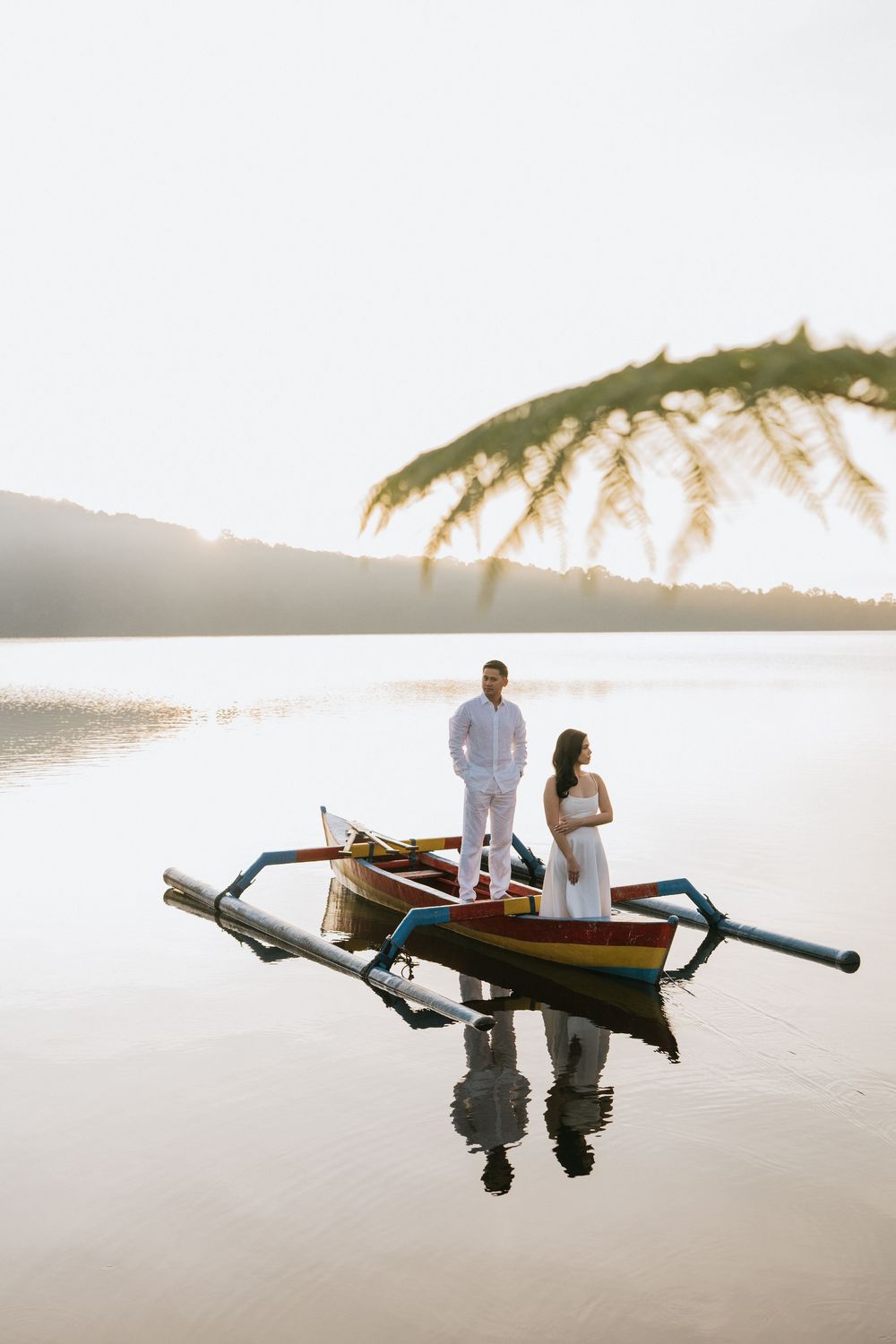 A wooden outrigger boat floats on mirror-like waters during golden hour with palm trees in the distance.
