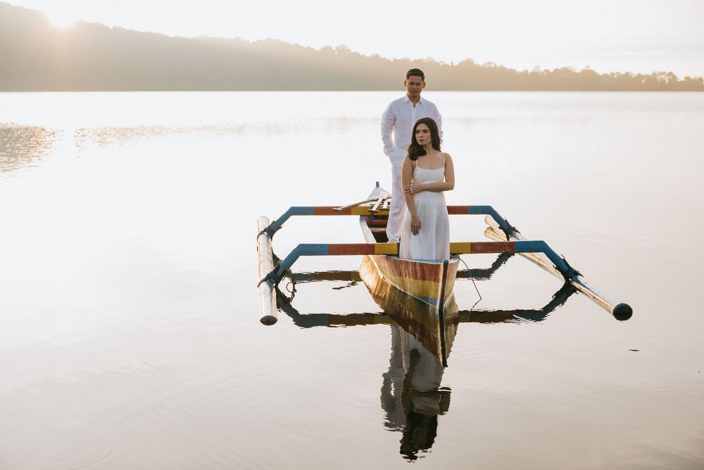 A couple sits in a traditional wooden boat on tranquil waters as the sun sets behind distant hills.