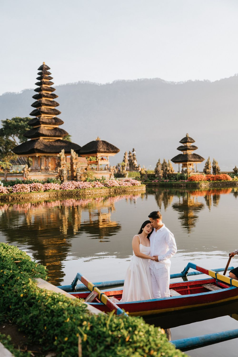 Couple in boat at Ulun Danu Beratan temple with mountain backdrop and reflective waters.