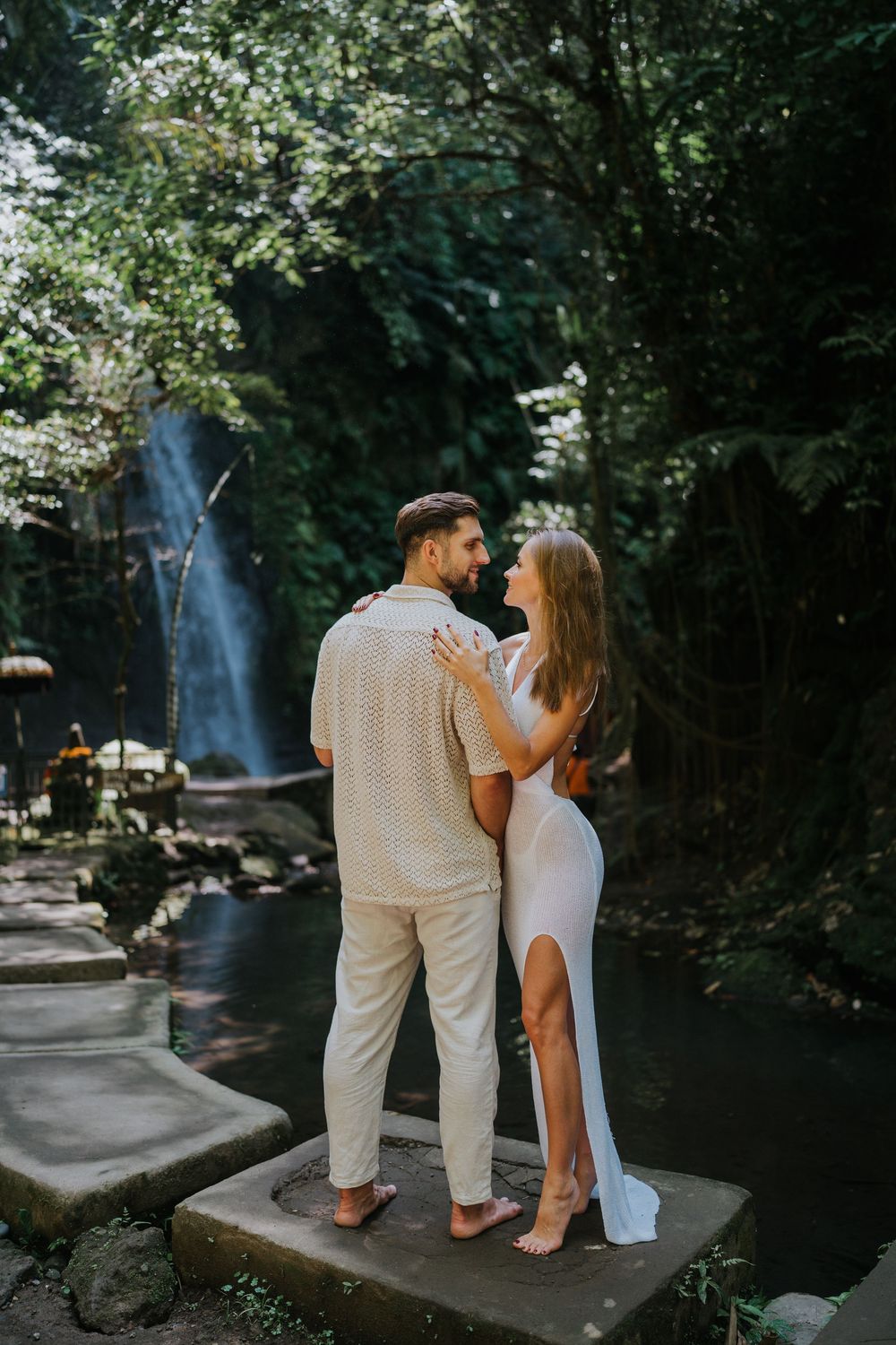 A couple in light colored outfits share an intimate moment by a waterfall in a lush forest setting.