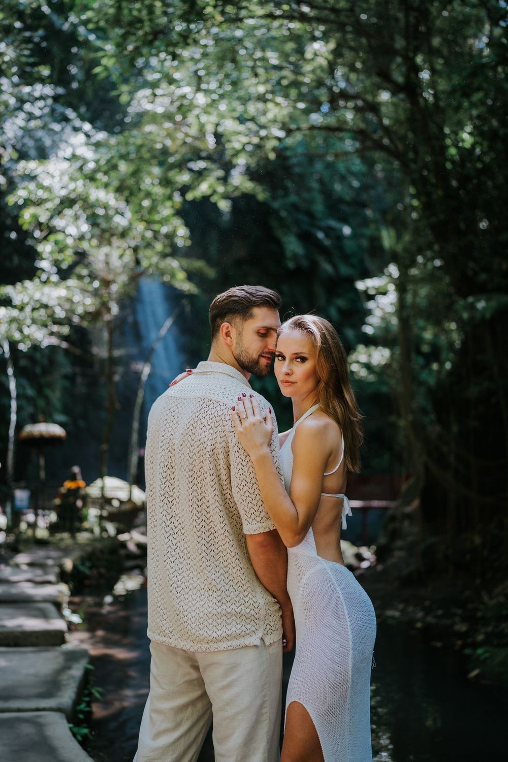 A romantic couple stands together against a backdrop of green foliage in matching light-colored attire.