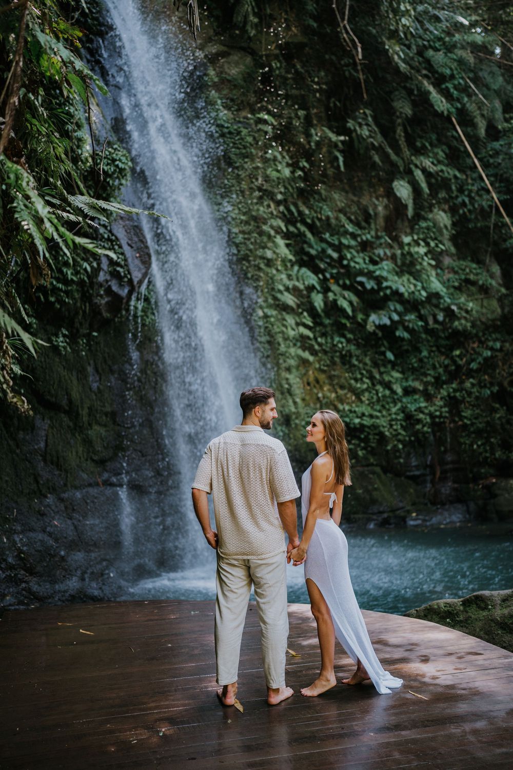 Two people in white clothing pose together near a cascading waterfall surrounded by tropical greenery.