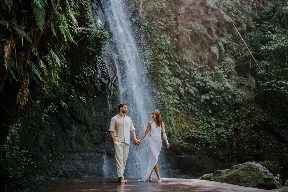 A couple stands on a wooden platform overlooking a scenic waterfall in a tropical forest setting.