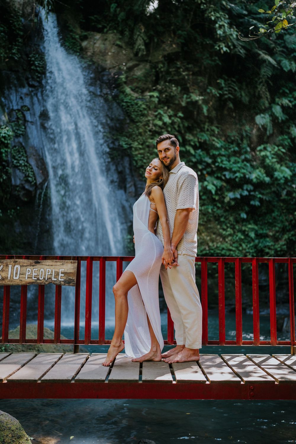 A couple in white outfits embraces on a red railing bridge near a tropical waterfall.
